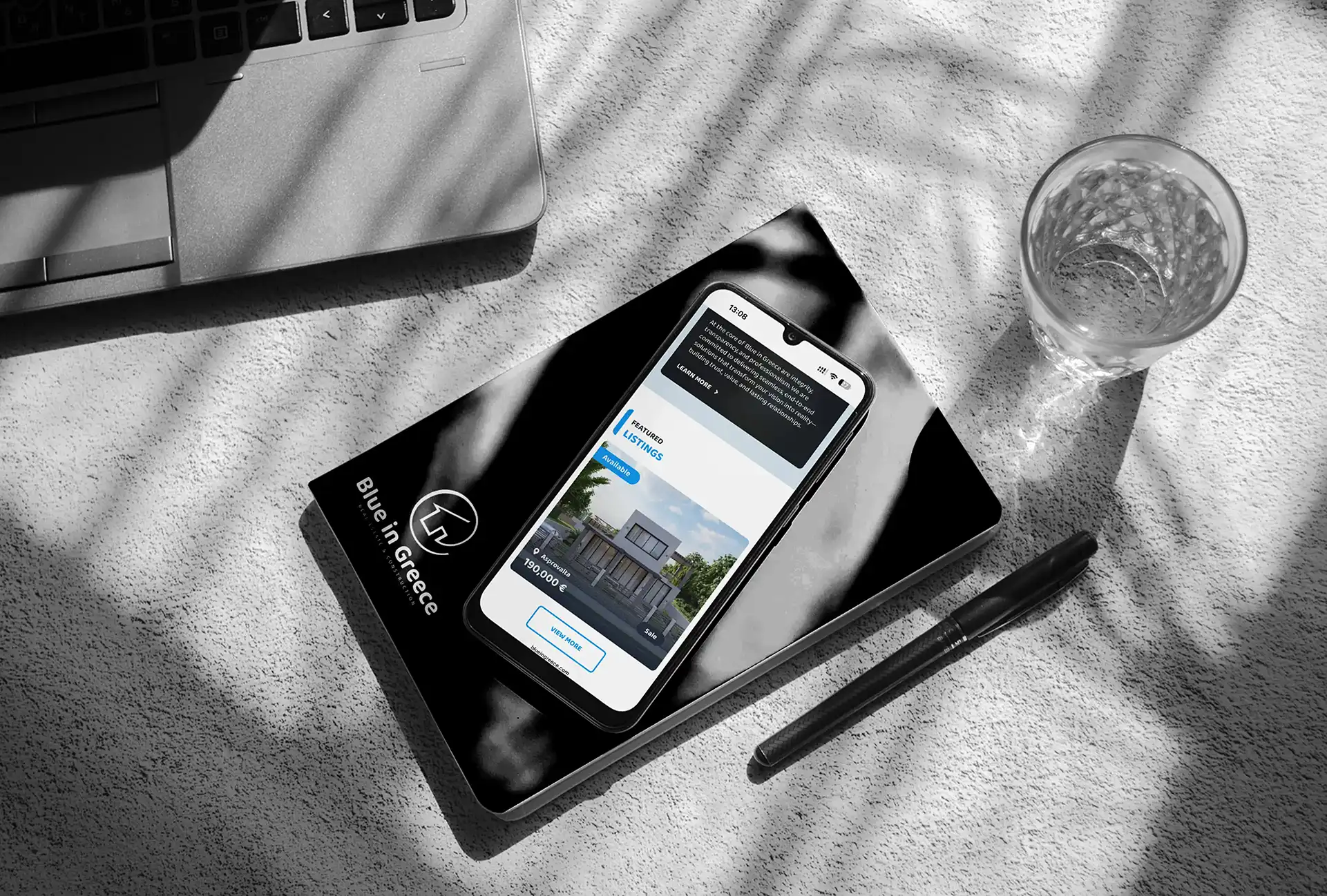 The Blue in Greece mobile site on a phone resting on a branded card, on a sunlit grey desk