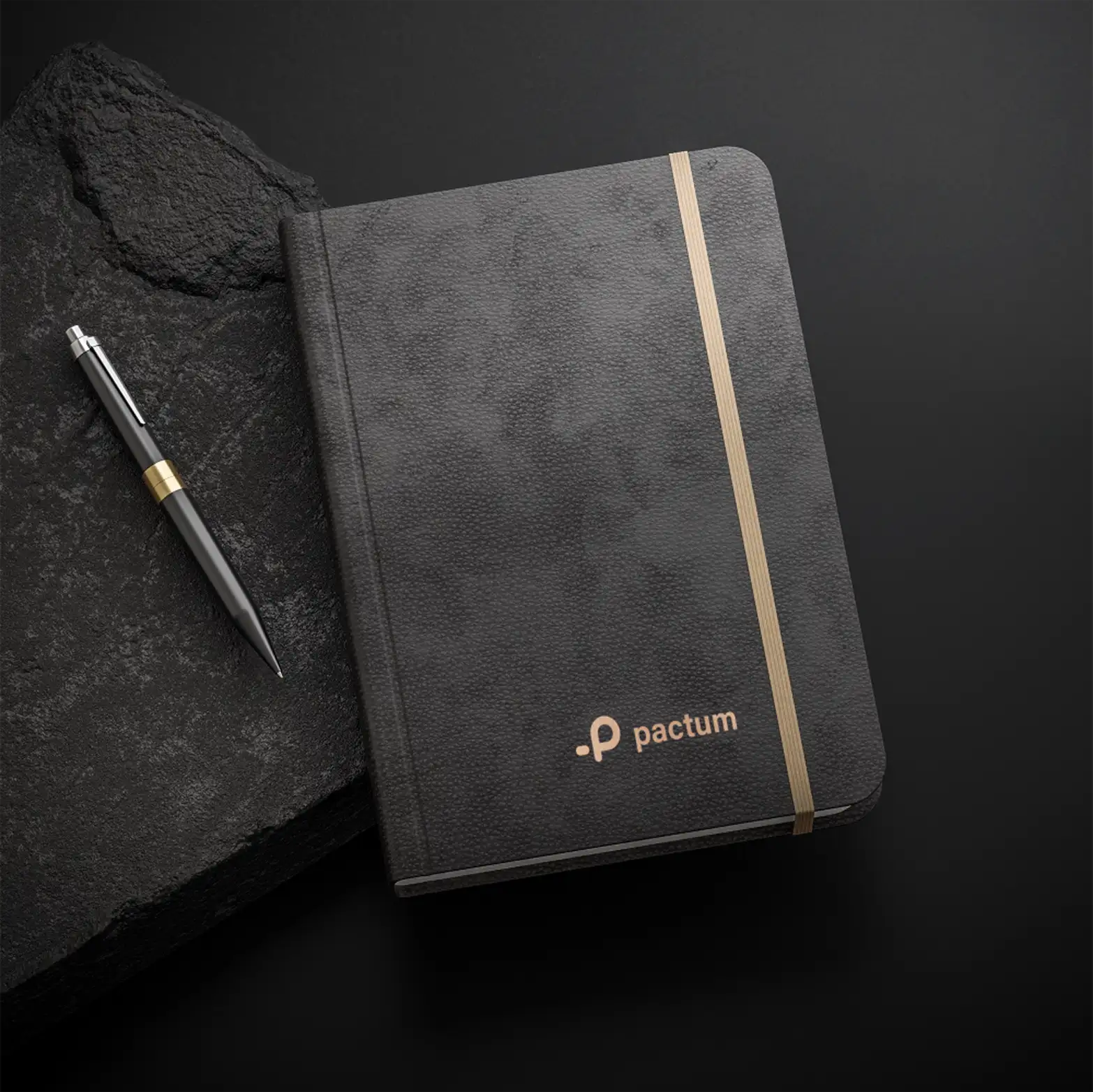 A black leather Pactum notebook with a gold foil logo and a matching pen on dark stone